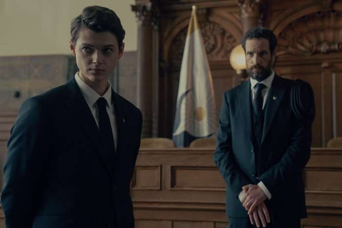 BRAD ALEXANDER, CHARLIE CARRICK Brad Alexander and Charlie Carrick in The Testaments standing at the front of a courthouse, wearing suits and looking serious down the room.