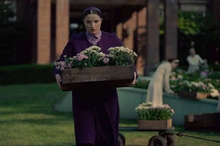LUCY HALLIDAY Lucy Halliday in The Testaments holding a planter of flowers, looking serious as she sneaks away into the garden, wearing a purple uniform and headband.
