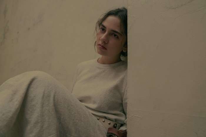 MATTEA CONFORTI Mattea Conforti in The Testaments sitting down in a prison cell, wearing a white gown and restraints, looking blankly at the other side of the room.