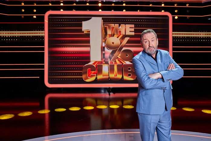 Lee Mack wearing a blue shirt and black shirt, stood with his arms folded.