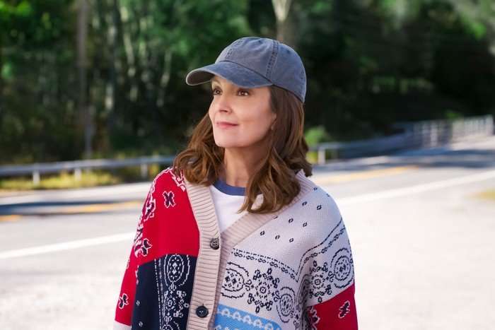 THE FOUR SEASONS, SEASON 2 A woman with shoulder-length brown hair stands beside a quiet road lined with trees, looking off into the distance with a slight smile. She is wearing a blue baseball cap, a white T-shirt and a colourful patterned cardigan in red, cream and blue tones. The background is softly blurred, with sunlight filtering through the wooded area behind her.