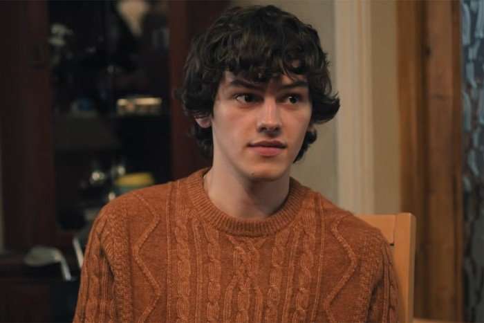 Jackson Connor stars in Tip Toe; in this scene, he is wearing an orange jumper and glancing over at someone with a slight smirk