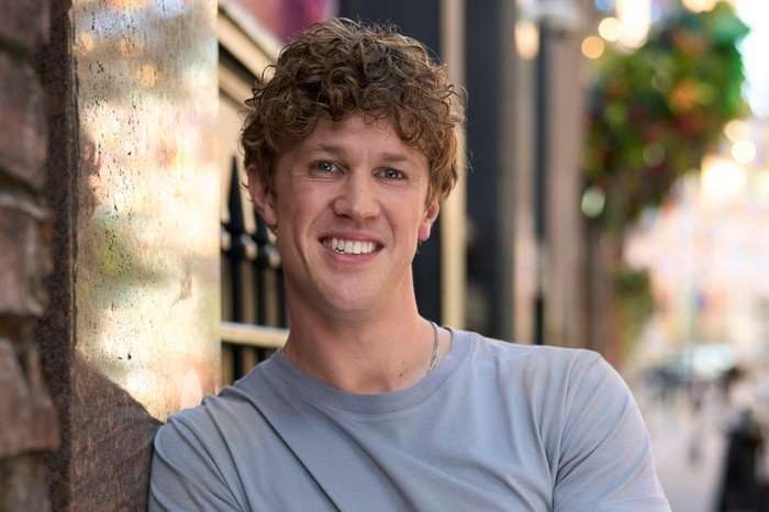 Joseph Evans as Saul in Tip Toe. He is leaning against an exterior wall, smiling on a bright day.