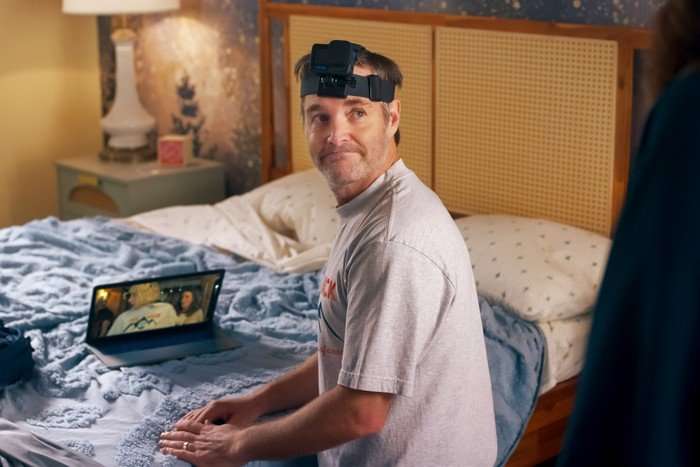 THE FOUR SEASONS, SEASON 2 A man sits on the edge of a bed in a warmly lit bedroom, looking over his shoulder with a resigned expression. He is wearing a grey T-shirt and a head-mounted camera strapped around his forehead. A laptop rests open on the bed beside him, while a bedside lamp and patterned wallpaper add to the cosy setting. Another figure is partially visible in the foreground on the right-hand side of the image.