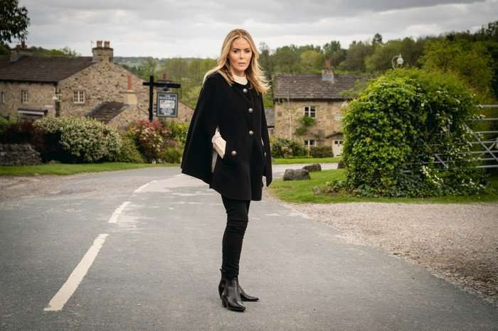 Sadie King, in a black cloak and black trousers, stood on the road to Emmerdale. She is played by Patsy Kensit.