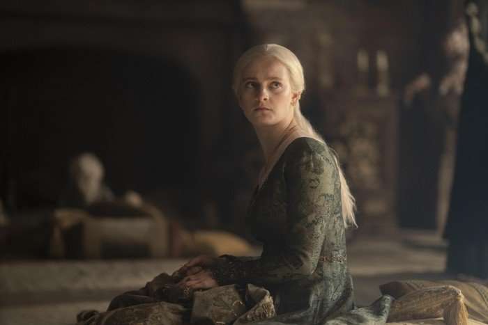 House of the Dragon Phia Saban sits and turns on a bed in a green dress as Queen Helaena Targaryen in House of the Dragon season 2.