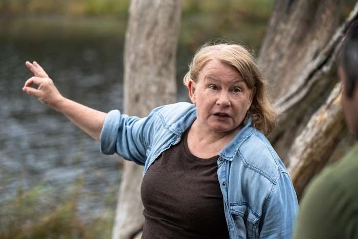 Donna stands by a river, gesturing with her hand as she explains something with a concerned look on her face