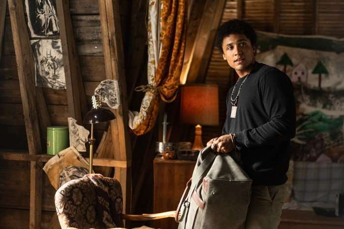 Ellis stands in a room decorated with shabby antique furniture, holding a large bag and looking off to the side