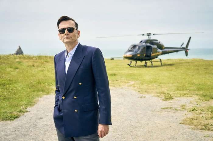 RIVALS 2 David Tennant in season 2 of Rivals. He is outside with a helicopter in the background; he wears aviator sunglasses and a double-breasted jacket