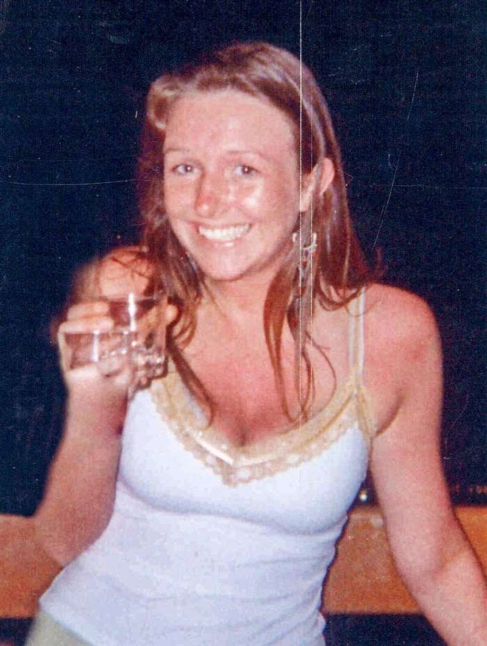 A young woman with long dark-blond hair in a white camisole top holding a glass