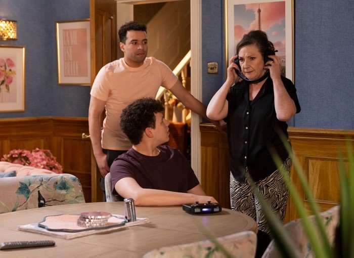 25_06_coro_will_maggie_03 Ollie and Will look at Maggie, who is wearing a pair of headphones, in a scene from Coronation Street