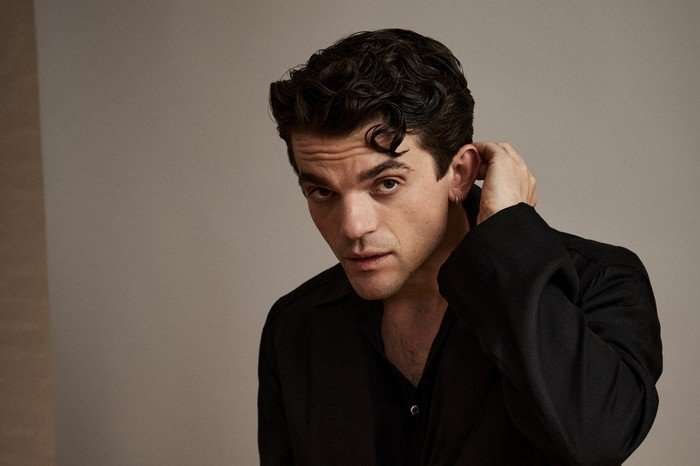 Edward Bluemel, wearing a black shirt and jacket and with his hand to his ear.
