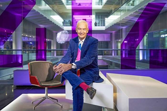 Jon Snow sitting crosslegged in a news studio, wearing a navy suit, colourful tie and red socks.