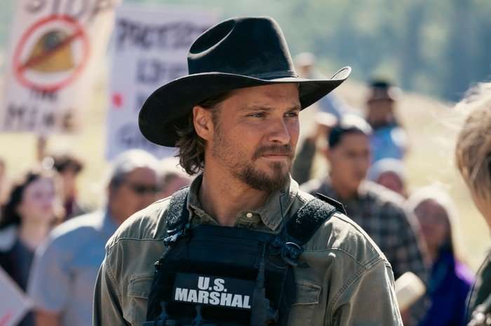 3098759_02764b Luke Grimes as Kayce Dutton in Marshals, wearing a vest with US Marshal written on it, along with a stetson, and stood in front of a crowd of people.