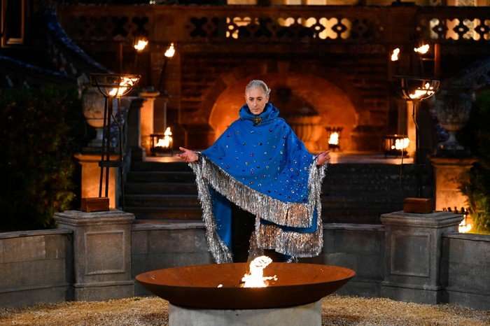 Alan Cumming stood outside Ardross Castle in front of a firepit, wearing a blue shawl with his arms stretched out.