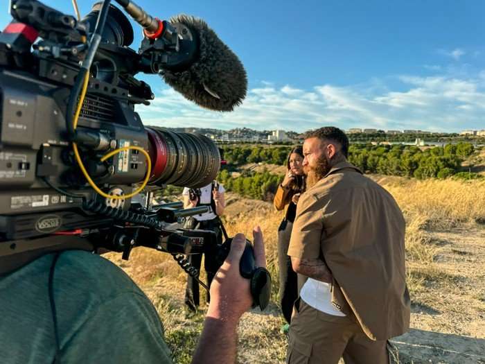 514486,TITLE:Ashley Cain: Into the Danger Zone A cameraman with a camera rested on his shoulder looking at Anais Merad and Ashley Cain.