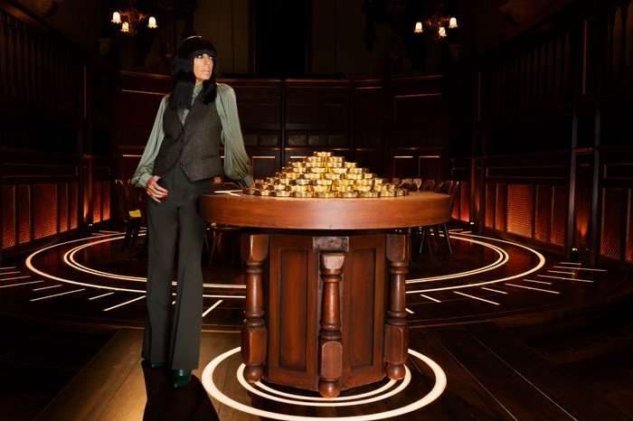 546795,TITLE:The Traitors S4 Claudia Winkleman stood next to a lectern filled with golden discs.