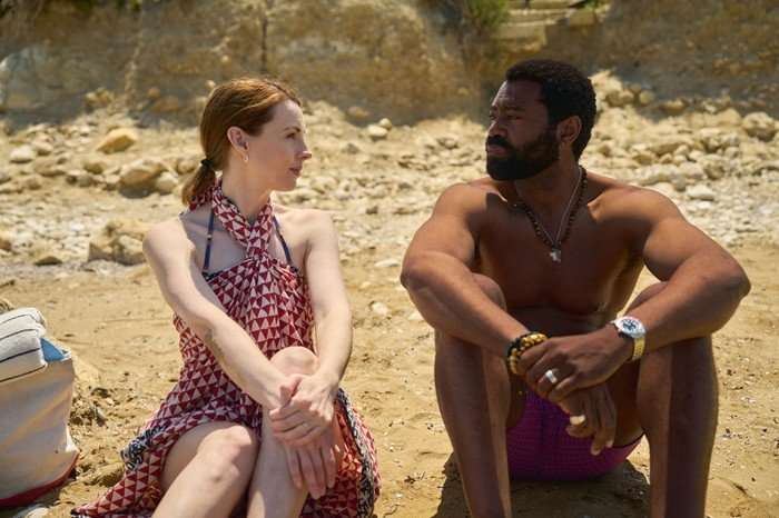 561474,TITLE:Two Weeks In August Jessica Raine as Zoe and Nicholas Pinnock as Solomon in Two Weeks in August, sat on a beach, looking at one another. She is wearing a red and white check dress and he is wearing swim shorts.