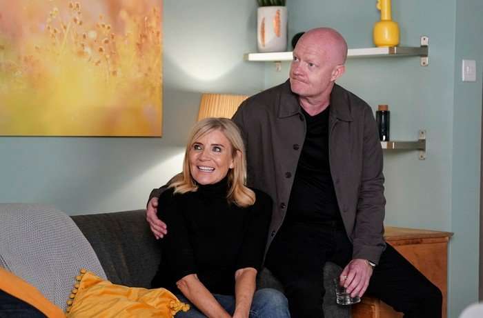Michelle Collins smiles as Cindy Beale sat on a sofa with Jake Wood as Max Branning with his hand around her as they look to the left of the frame in EastEnders.