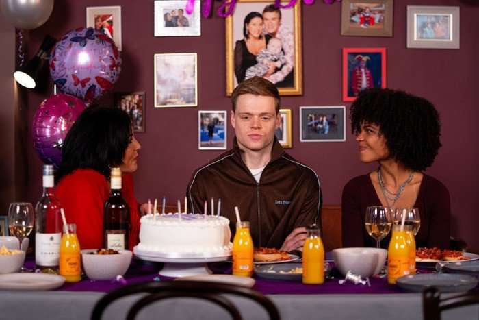 564107,TITLE:Eastenders Oscar sits awkwardly between Kat and Jasmine, with a birthday cake in front of him, in a scene from EastEnders