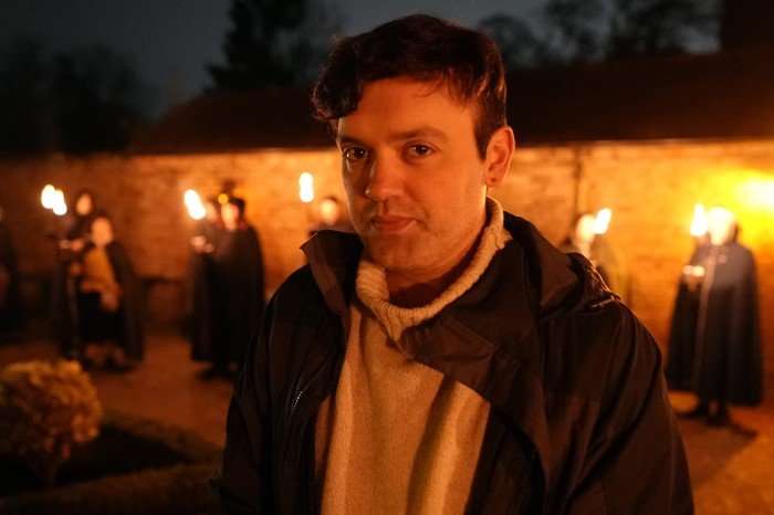 567855,TITLE:Queen James Gareth Russell stood in a courtyard with people behind him holding lanterns as he looks ahead.