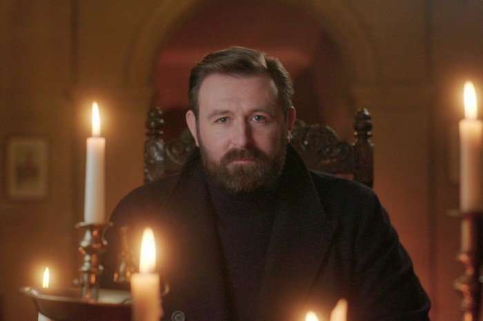567857,TITLE:Queen James James McArdle sat in a throne chair with candles around him, looking ahead.