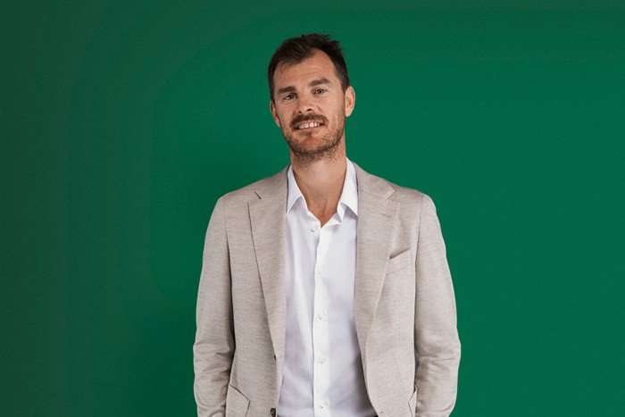 Ex-tennis player Jamie Murray, in a grey jacket and white shirt, stands smiling in front of a green background.