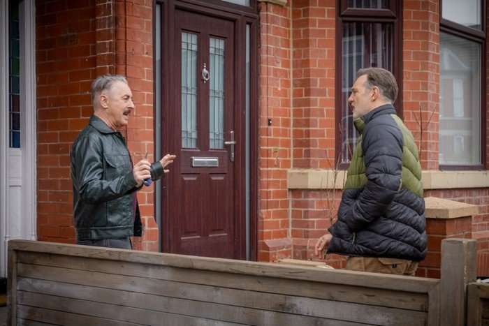Alan Cumming and David Morrissey in Tip Toe. Cumming's character wears a leather jacket and has a defensive stance as he talks to his neighbour outside the front door. Morrissey's character wears a dark blue and green puffer jacket.