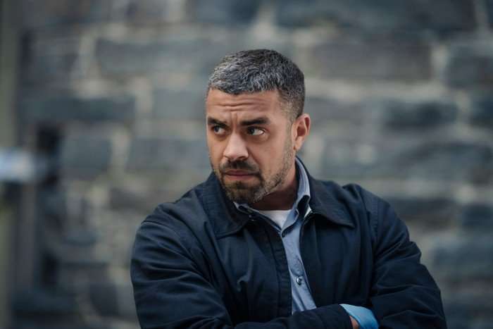 78256_1_S2_Ep1_- Embargo 0001 Tuesday 30th December - Patience Series 2 - Ep1 Nathan Welsh plays DS Jake Hunter in Patience season 2; he is seen here wearing a jacket and shirt, standing before a blurred out stone brick background