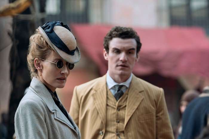 Natascha McElhone as Cordelia Holmes and Dónal Finn as James Moriarty in Young Sherlock. He is wearing a suit and she is wearing a jacket, hat and sunglasses.