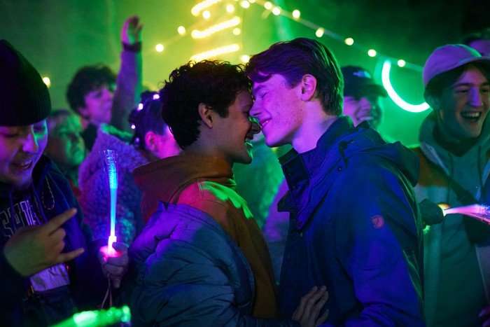 Young Royals season 3 Simon and Prince Wilhelm at a party, with a lively crowd around them and neon lights in the background