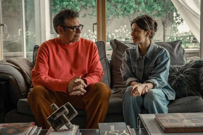 Jemaine Clement and Yali Topol Margalith in Alice and Steve; in this scene, Steve and Izzy are sat next to each other on a grey sofa in his stylish, modern living room, with a bright window behind them