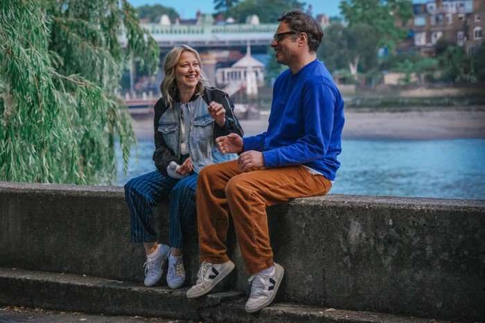 Nicola Walker and Jemaine Clement in Alice and Steve, sat on a wall by a river laughing together.