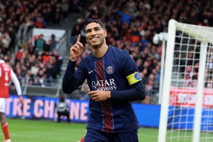 Achraf Hakimi Achraf Hakimi, in blue and red PSG football kit, smiles and holds his index finger up as he celebrates scoring.