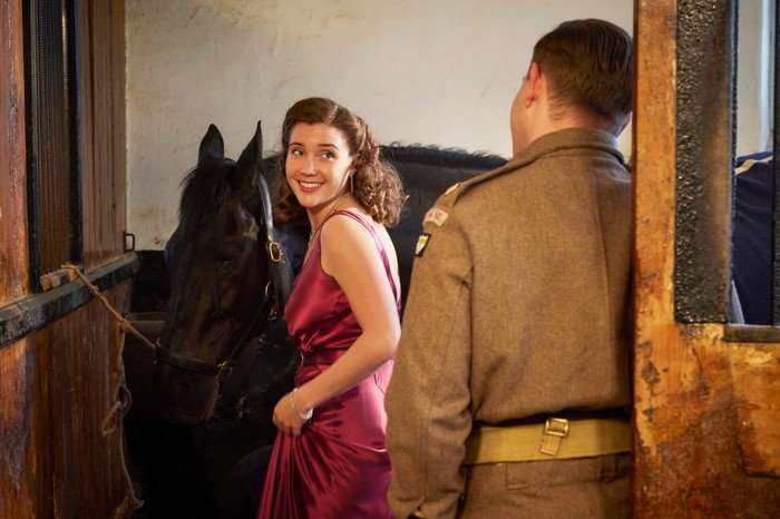 Gaia Wise as Charlotte Beauvoir, stood next to a horse in a stable wearing a long red dress, looking over her shoulder and smiling at Callum Woodhouse as Tristan, who is dressed in a military uniform