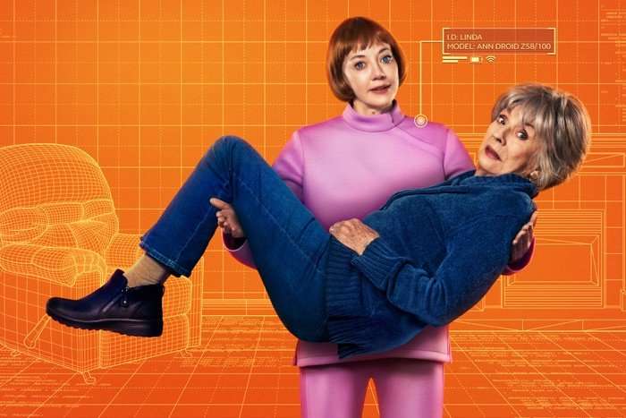 AnnDroid-KEYART_568798 Diane Morgan and Sue Johnston in Ann Droid. Morgan is dressed in pink and carrying a worried looking Johnston, who is wearing blue. They are against an orange backdrop.