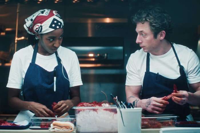 Jeremy Allen White and Ayo Edebiri in The Bear, chopping food and looking at each other in the kitchen, while wearing white t-shirts and blue aprons.