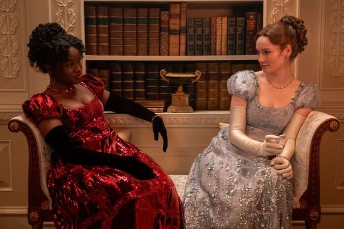 Bridgerton-s4-part2-Masali-Hannah Masali Baduza as Michaela, Hannah Dodd as Francesca Bridgerton in Bridgerton sitting on a sofa wearing bedazzled ball gowns and staring at each other.