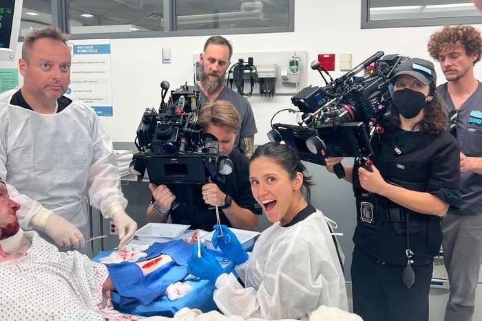 Credit Melette Cabot Dr Elizabeth Ferreira behind the scenes of The Pitt, wearing a white covering and smiling at the camera. There is a camera crew stood behind her and she is holding a medical tool, knelt by an actor playing a patient, lying in a hospital bed.