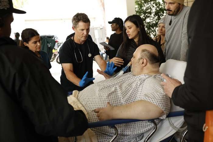 BTS Dr Elizabeth Ferreira behind the scenes of The Pitt, wearing black, stood next to an actor playing a patient, lying on a bed. Others are stood around, including Shawn Hatosy, who is wearing blue hospital scrubs and latex gloves.