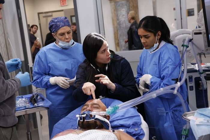 BTS Dr Elizabeth Ferreira behind the scenes of The Pitt, wearing black, pointing towards an actor playing a patient, lying on a bed. Two other actors are stood either side of her, wearing blue hospital scrubs and face masks.