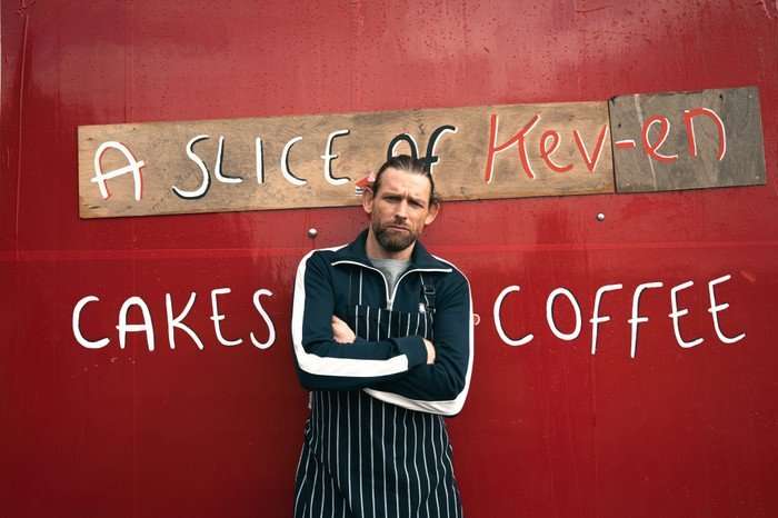 EMBARGO_EMM_KEV_BACK Chris Coghill as Kev Townsend poses against a red wall in an apron with the sign behind saying "A Slice of Kev-en...Cakes and Coffee' in Emmerdale.