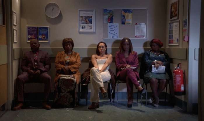 From left to right, Howie Danes, Yolande Trueman, Chelsea Fox, Denise Fox, and Kim Fox sit in a hospital waiting room with a clock on the wall reading both hands at 1 in EastEnders.