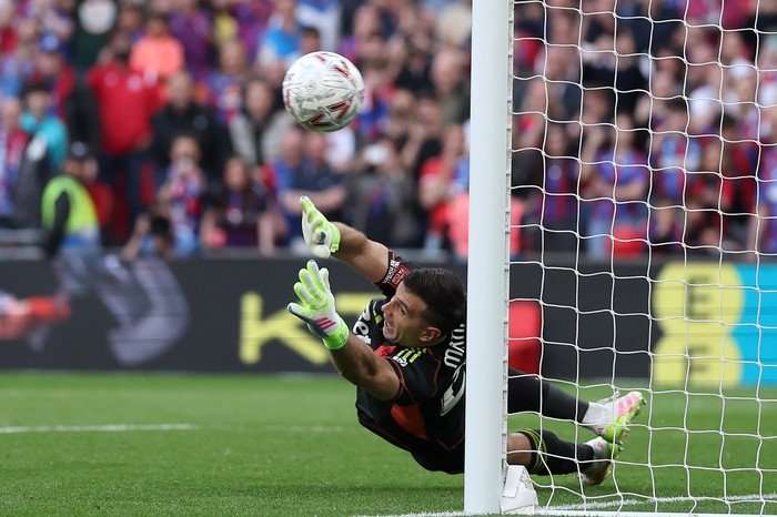 Emiliano Martinez, in black Aston Villa goalkeeping kit and green gloves, dives to make a penalty save with both hands.