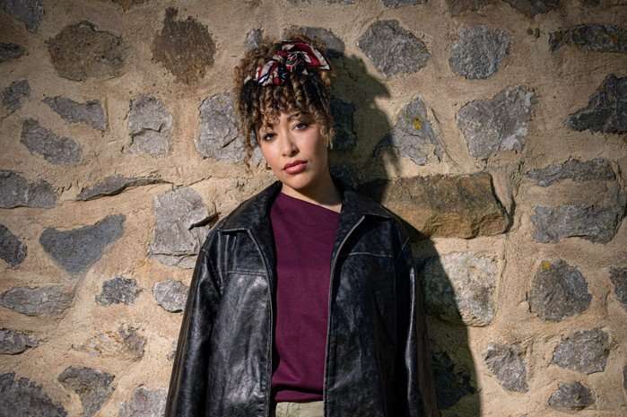 Casey Al-Shaqsy as Serena Sugden in Emmerdale, stood against a stone wall and wearing a black leather jacket.