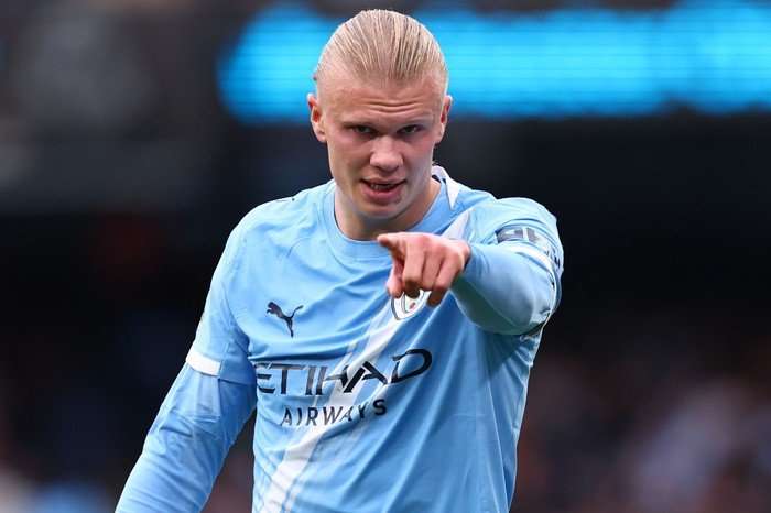 Erling Haaland, in a light blue Man City football shirt, points at the camera with his left hand.