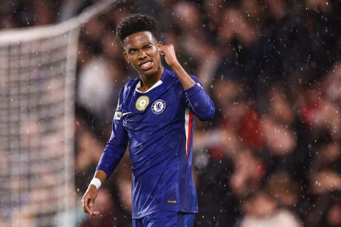 Estevao, in blue Chelsea football kit, smiles and winks as he holds his right fist up in celebration in the rain.