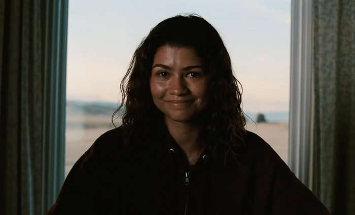Zendaya smiles while seated in a hoodie as Rue Bennett in Euphoria season 3 episode 8.