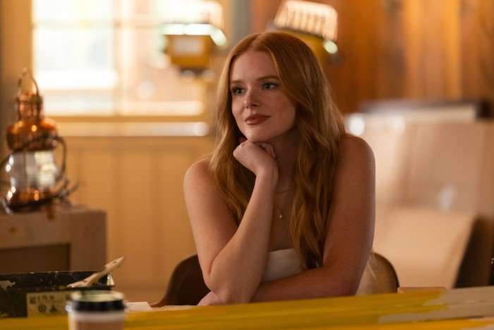 Every Year After - First Look Abigail Cowen as Delilah sitting in a room, with sunlight coming in and smiling as she rests her head on her hand.