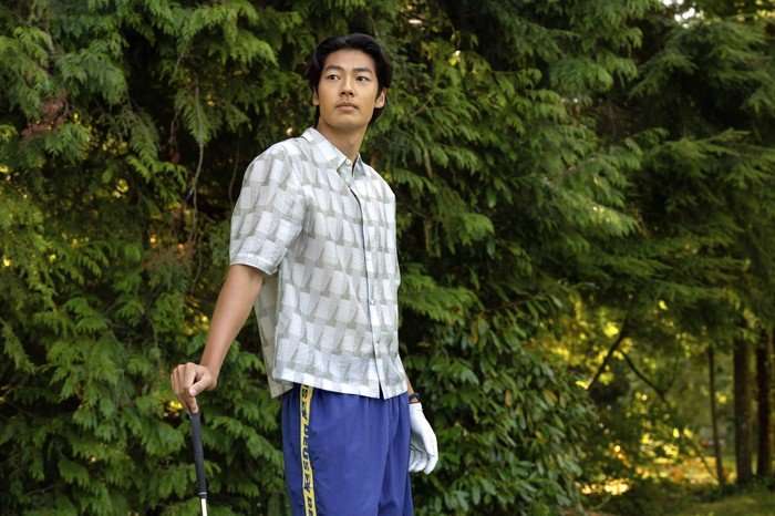 Every Year After Joseph Chiu as Jordi standing against his golf club in a patterned shirt and blue sports shorts.